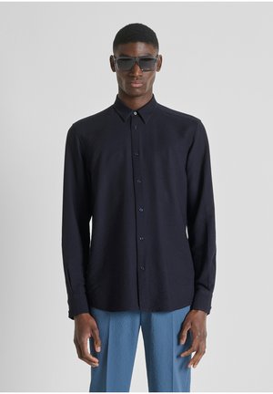 Hombre vestido con una camisa de botones azul marino oscuro, pantalones texturizados azul claro y gafas de sol oscuras, de pie frente a un fondo claro y liso.