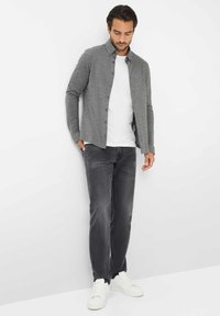 Graues Hemd über einem weißen T-Shirt, kombiniert mit dunkelgrauen Jeans und weißen Sneakern. Glatte Textur, taillierte Passform, lässiger Stil.