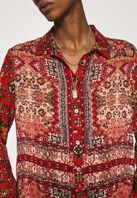 Blouse rouge à motifs avec des designs complexes en noir, rose et or. Comprend un col, une fermeture à boutons devant et des manches longues.