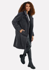 Grauer Kunstfellmantel mit lässiger Passform, Seitentaschen und Reverskragen, kombiniert mit schwarzen Lederleggings und Stiefeletten.