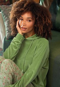 Vrouw met krullend haar draagt een groene hoodie en een gebloemd broek, zit comfortabel binnen met haar hand op haar gezicht.