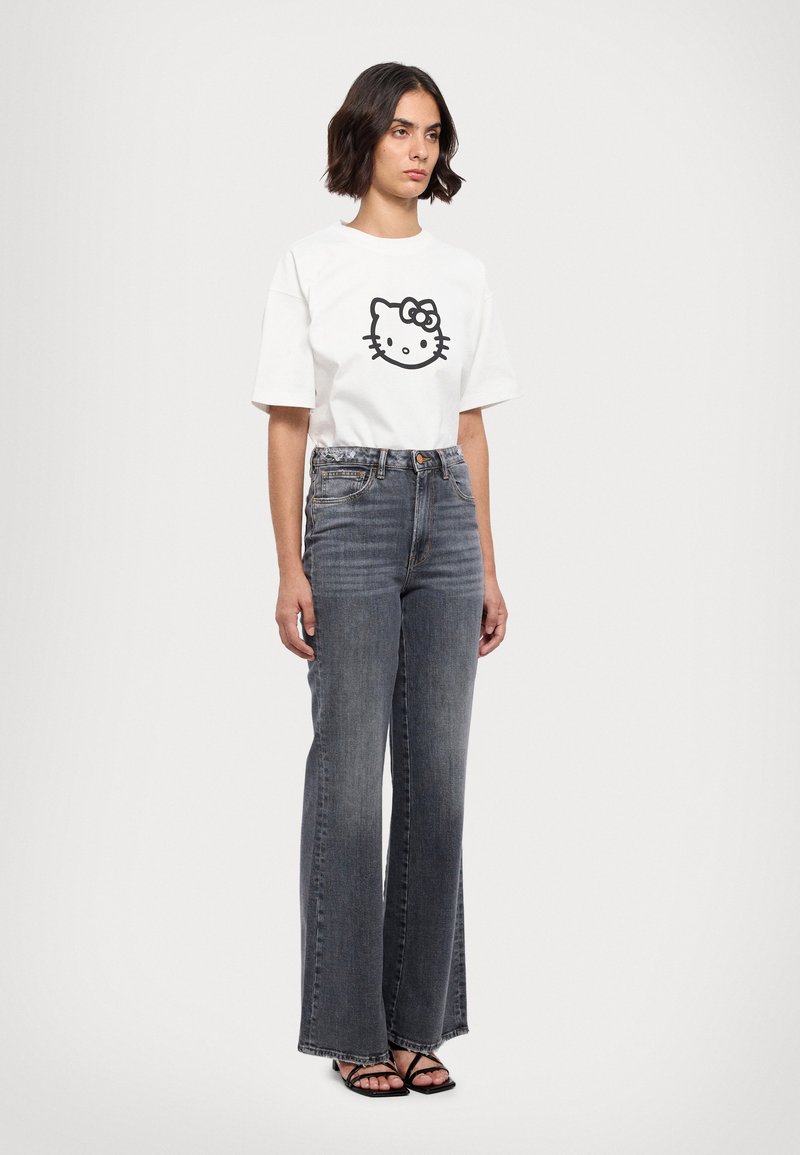 Tricou alb din bumbac cu un grafic mare în negru cu Hello Kitty, asociat cu blugi din denim gri cu talie înaltă și sandale negre cu barete.