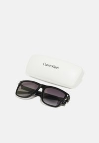 Črne pravokotne sončne očala z temno gradacijo leč ob belem trdem ovitku blagovne znamke Calvin Klein na belem ozadju.