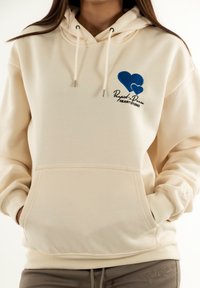 Sudadera cremada con capucha y bordado de corazón azul, con un bolsillo delantero y cordones blancos. Textura suave, corte casual.