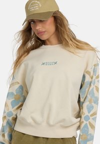 Sudadera color crema con mangas de patrón floral azul, escote redondo y puños acanalados, combinada con una gorra beige con texto bordado.