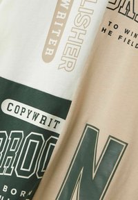 Dos camisas de algodón plegadas con texto impreso en beige, crema, verde oscuro y blanco, que presentan palabras como "COPYWRITER" y "PARA GANAR EL CAMPO."