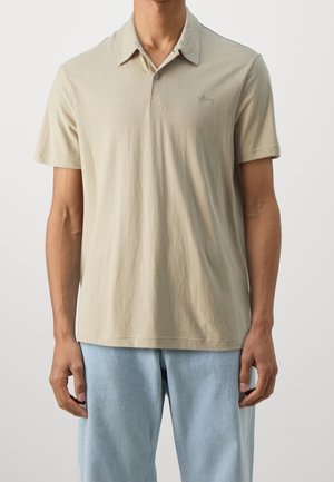 Mężczyzna w beżowej koszulce polo z krótkim rękawem i jasnoniebieskich dżinsach, stojący na tle jednolitego jasnego tła.