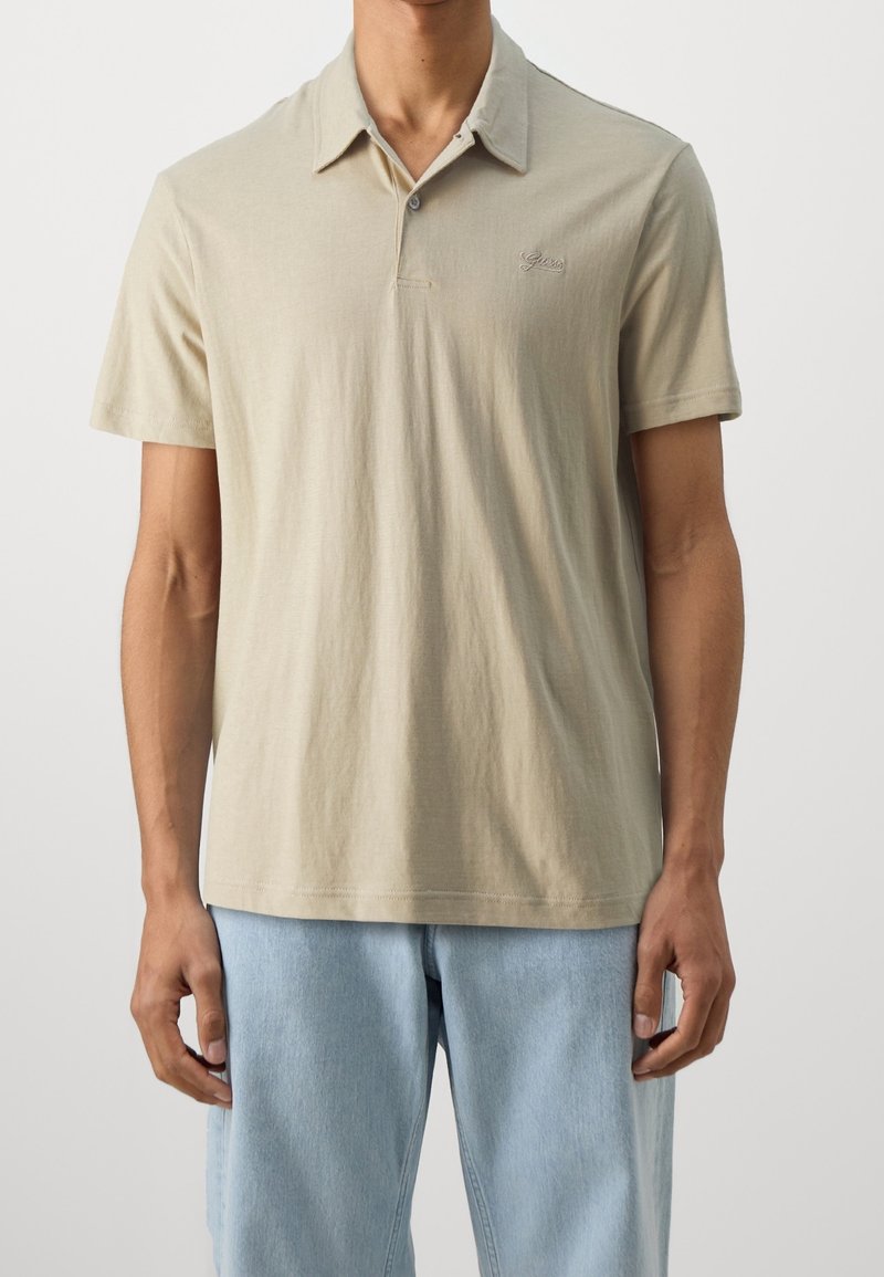 Homme portant un polo beige à manches courtes et un jean bleu clair, debout devant un fond clair uni.