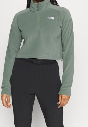 Woman wearing a cropped green quarter-zip fleece with The North Face logo and black athletic pants with a mesh waistband.