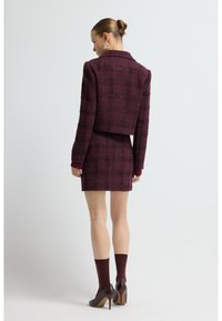 Ensemble blazer en tweed à carreaux bordeaux et jupe mini, mettant en avant un tissu texturé, un design ajusté et des accents bordeaux contrastants sur les manches et les chaussettes.
