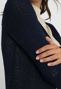 Mörkblå, stickad cardigan med ett löst, öppet vävmönster. Har ett mjukt, texturerat tyg och en avslappnad passform. Ses bäras över en ljus topp.