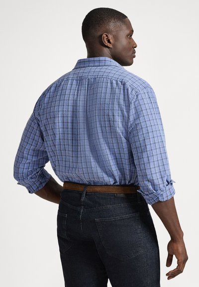 Modro karo srajca s pritrdilnimi gumbi in zavihki na rokavih; izdelana iz lahkega blaga. Oblečena čez temno denim jeans hlače, s rjavim pasom.