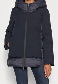 Námořnická puffer bunda s velkou kapucí, zipovým zapínáním, polstrovanou dolní částí a bočními kapsami. Obsahuje kontrastní textury a materiály.