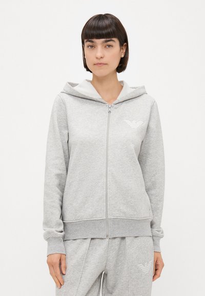 Haut à zip gris clair fabriqué en matériau doux, avec un logo blanc et une capuche contrastante. Poignets et taille côtelés.
