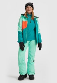 Skikleding met een turquoise en oranje jas, turquoise basislaag en muntgroene broek. Bevat zwarte handschoenen en skibril.