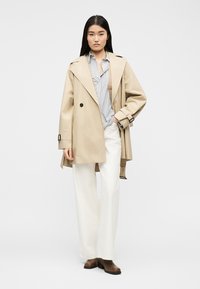 Γυναίκα που φοράει μπεζ καμπαρντίνα, ριγέ πουκάμισο, λευκό παντελόνι φαρδύ και καφέ μπότες, στέκεται με ουδέτερη έκφραση.