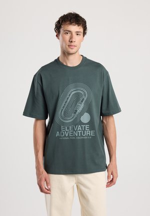 Dunkelgrünes Baumwoll-T-Shirt mit kurzen Ärmeln, das ein helles grafisches Design mit dem Text „ELEVATE ADVENTURE“ und einem kreisförmigen Muster in der Mitte zeigt.