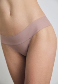 Lichtroze naadloze stringonderbroek met een gladde textuur en een brede tailleband, voorzien van subtiele branding. Het materiaal lijkt rekbaar en comfortabel.