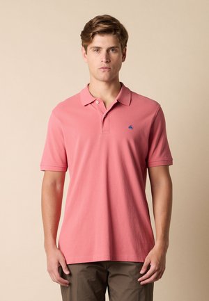 Junger Mann mit kurzen hellbraunen Haaren, der ein rosa kurzärmeliges Polohemd und braune Hosen trägt, steht vor einem beigen Hintergrund.
