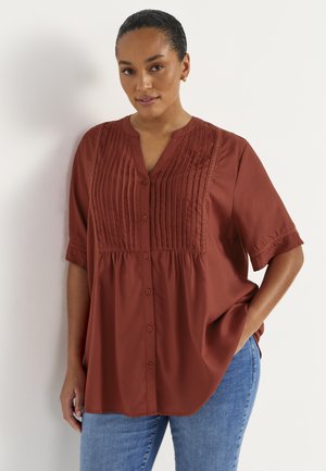 Kvinde med mørkt hår trukket tilbage iført en løs rustfarvet knapbluse med folder og blå jeans, stående op ad en hvid væg.