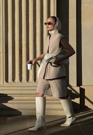 Beige ärmlös ytterplagg över en ljusbeige klänning, matchad med knähöga vita stövlar. Accessoarerna inkluderar solglasögon, en huvudduk och en kaffekopp.