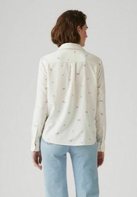 Camicia bianca con stampa floreale in vari colori, dotata di colletto a punta, maniche lunghe e orlo leggermente curvato.