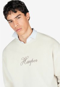Felpa beige realizzata in morbido tessuto con collo rotondo, caratterizzata da un ricamo in corsivo grigio della parola "Harper" sul petto.