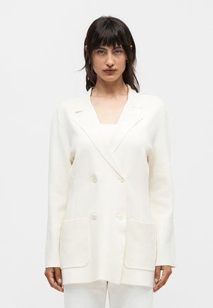 Femme aux cheveux foncés portant un blazer blanc croisé avec poches avant, debout devant un fond clair uni.