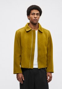 Jarek rumene gorčice z zadrgo spredaj, širokim ovratnikom in teksturirano tkanino. Ima preproste linije in sproščen kroj. Nošen čez belo srajco.