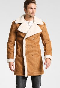 Hombre con cabello corto que lleva un abrigo de shearling de doble botonadura marrón con forro de piel blanca en el cuello, los puños y los bordes frontales.