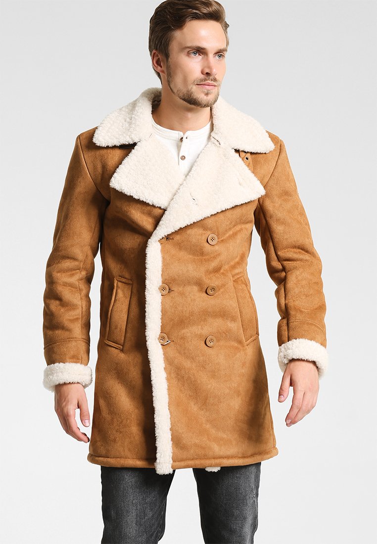 Hombre con cabello corto que lleva un abrigo de shearling de doble botonadura marrón con forro de piel blanca en el cuello, los puños y los bordes frontales.