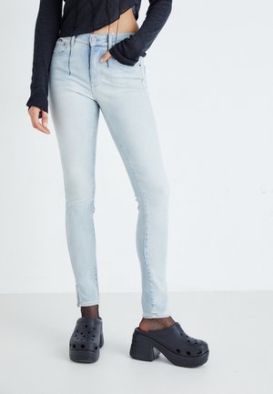 Persoon draagt lichtblauwe skinny jeans, zwarte chunky plateauklompen en een kort zwart langärmelig shirt met duimgaten.