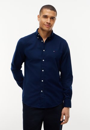 Homme aux cheveux courts portant une chemise bleu marine boutonnée, debout devant un fond blanc uni, souriant légèrement.