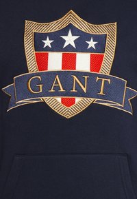 Sudadera navy con un gran diseño de escudo bordado que presenta rayas rojas y blancas, tres estrellas y "GANT" en letras doradas sobre un banner azul.