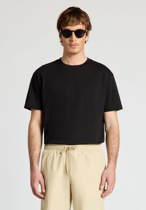 Junger Mann mit schwarzem T-Shirt, beigen Kordelhosen und runden schwarzen Sonnenbrillen steht vor einem einfarbigen Hintergrund.