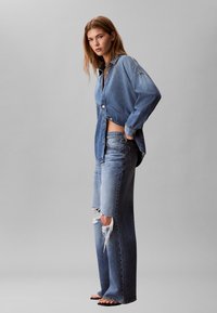 Junge Frau steht seitlich und posiert vor einem schlichten grauen Hintergrund, trägt ein blaues Jeanshemd, zerrissene, weit geschnittene Jeans und Sandalen.