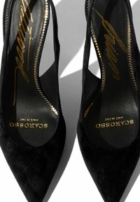 Crne suede cipele na visoku petu s oštrim vrhovima, koje imaju meku teksturu i zlatni logo na ulošcima uz detaljno šivanje.