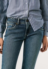 Modré džíny s slim fit střihem, s knoflíkovým a zipovým zapínáním, pěti kapsami a jemně vybledlým texturováním. V kombinaci s pruhovanou košilí.