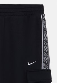 Juodi sportiniai kelnių, pasižymintys minkšta tekstūra, elastingu juosmeniu ir priešinga šonine juostele su kartotiniais "NIKE" tekstu ir logotipu.