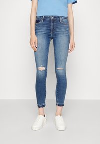 Vaqueros ajustados de mezclilla azul medio, con desgastes en las rodillas, un ligero desvanecimiento y dobladillos deshilachados, combinados con zapatillas blancas.