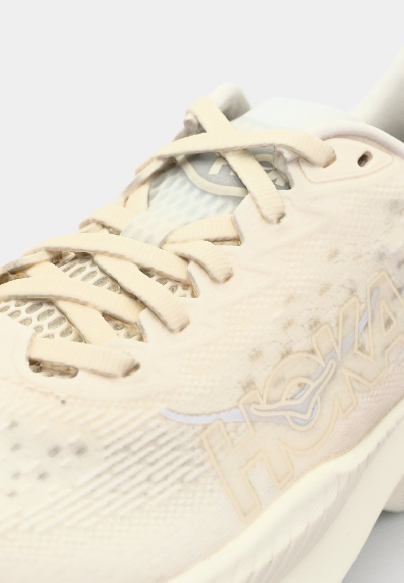 Jasno beżowy but sportowy z siateczkową górną częścią, sznurowaniem, logo HOKA w subtelnym żółtym odcieniu oraz teksturowaną podeszwą zapewniającą przyczepność.