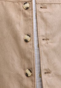 Gros plan d'une veste en daim beige avec des boutons marron clair et des boutonnières assorties, montrant partiellement une chemise blanche en dessous.