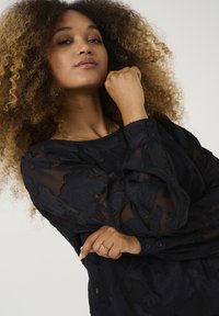 Blusa nera con motivo floreale, maniche lunghe trasparenti, tessuto texture, scollatura rotonda e polsini con bottoni. Il modello ha i capelli ricci e indossa un anello.