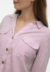 Camisa a rayas en rosa y blanco, con escote en V, dos bolsillos frontales y cierres de botones. Tejido suave con un corte relajado.