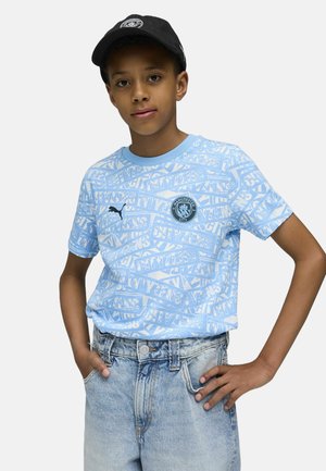 Maglietta grafica azzurro chiaro con testo bianco e logo del Manchester City, indossata con pantaloni corti in denim azzurro chiaro e un cappellino nero.