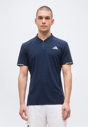 Mandlig model iført en marineblå kortærmet poloshirt med lynlåslukning ved kraven og hvide sportsshorts på en ensfarvet baggrund.