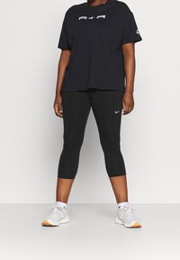 Camiseta de algodón negra con logo blanco, combinada con leggings capri negros que presentan un swoosh blanco de Nike, y zapatillas deportivas gris claro con acentos en beige.