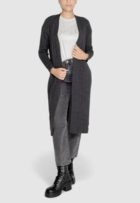 Cardigan lungo grigio scuro a coste con apertura frontale, abbinato a una maglietta chiara con design "Smile" e jeans neri a vita alta.