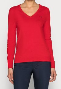 Pull rouge à col en V en tissu tricoté, avec manches longues et petit logo sur la poitrine. Associé à un pantalon foncé.
