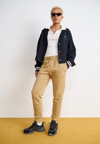 Melnā varsity jaka ar baltajām svītrām, balta polo krekla, bēšas sporta bikses ar Nike logotipu un melniem sporta apaviem. Teksturēta auduma, brīva forma.